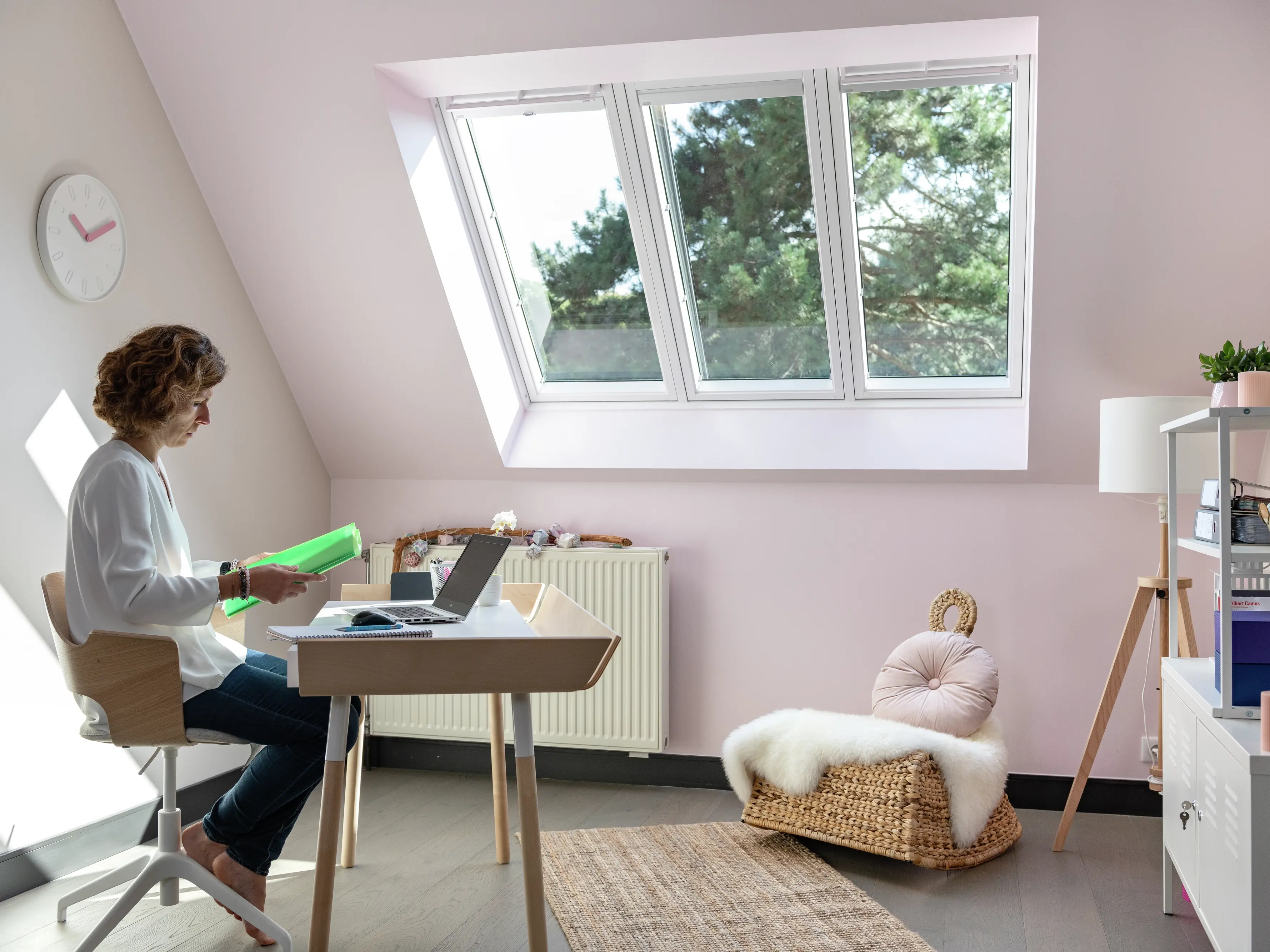 Une femme concentrée sur la lecture dans un bureau à domicile avec des murs roses, une fenêtre de toit, et un décor minimaliste comprenant une chaise tissée et des plantes vertes.