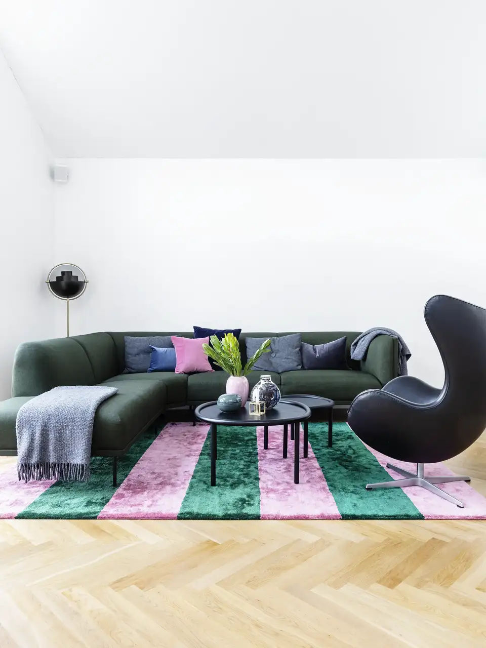 Salon rétro-pop avec canapé vert aux lignes douces, tapis géométrique rose et vert et fauteuil noir sculptural.