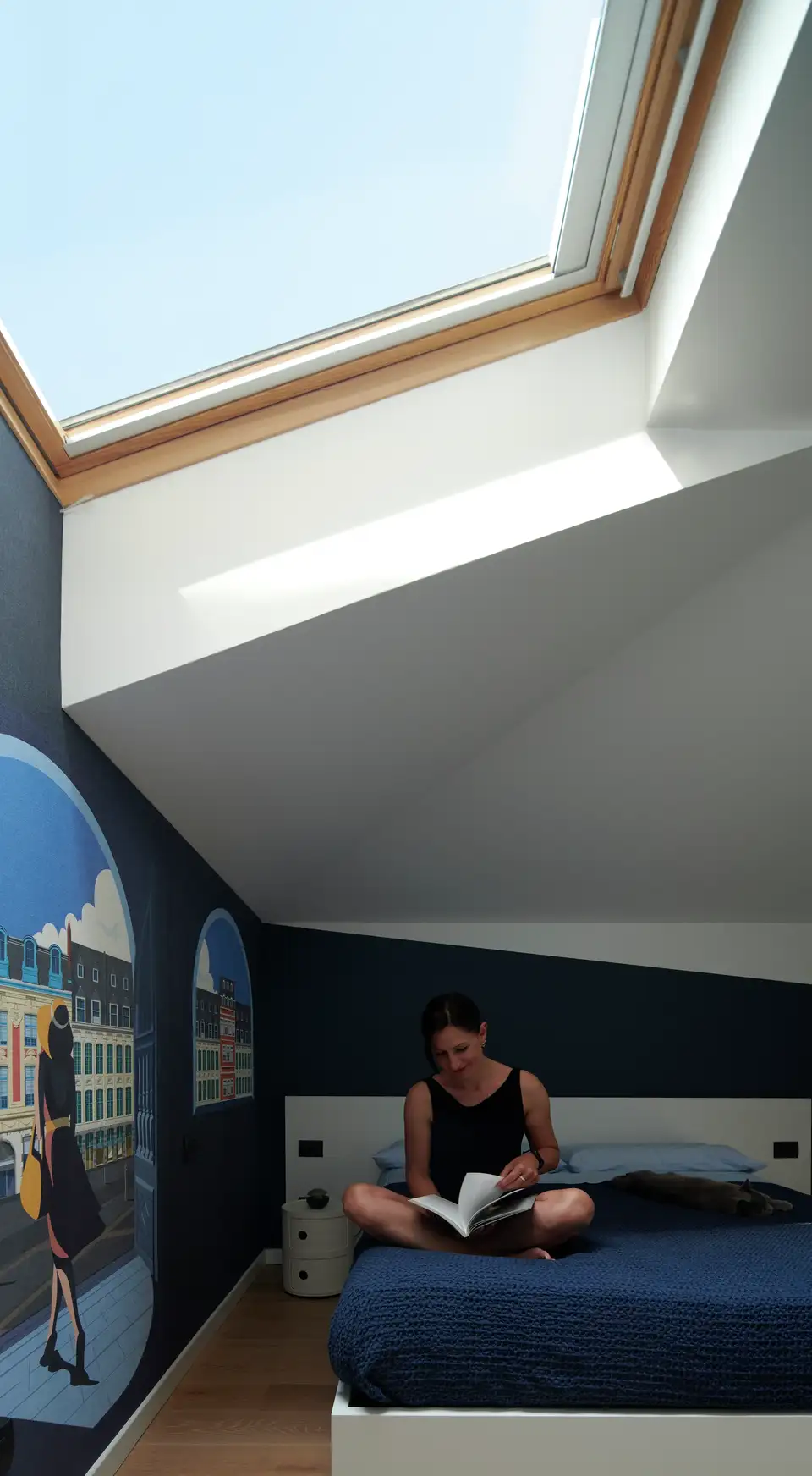 Kobieta siedzi na łóżku pod oknem dachowym, czytając książkę. Na ścianie za nią widnieje mural z miejskim krajobrazem.