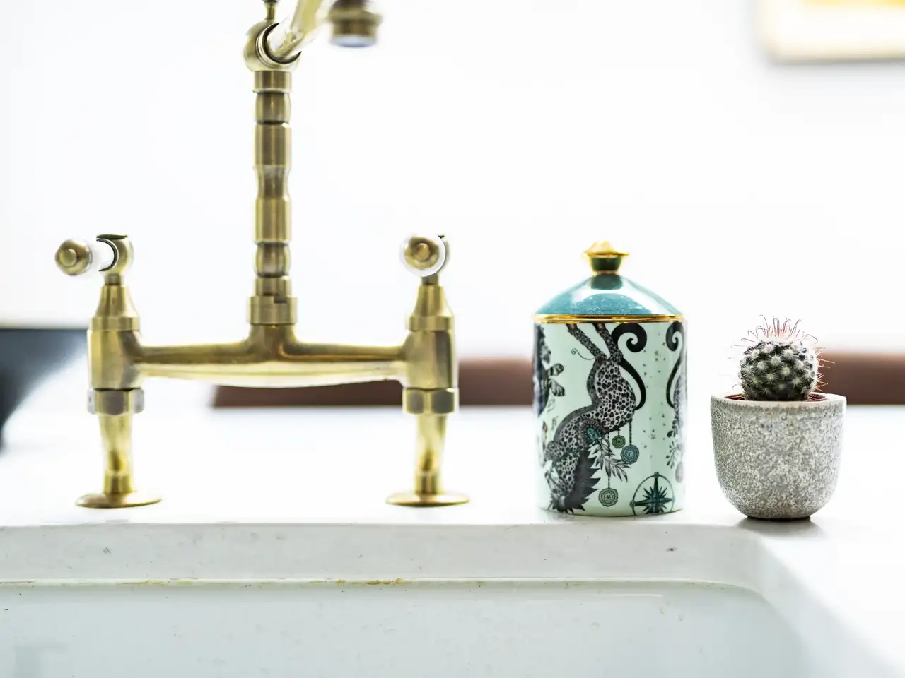Pirinç musluk, sanatsal desenlere sahip bir kavanoz ve dekoratif saksıda küçük bir kaktüs ile tamamlanan şık bir mutfak lavabosu düzenlemesi.