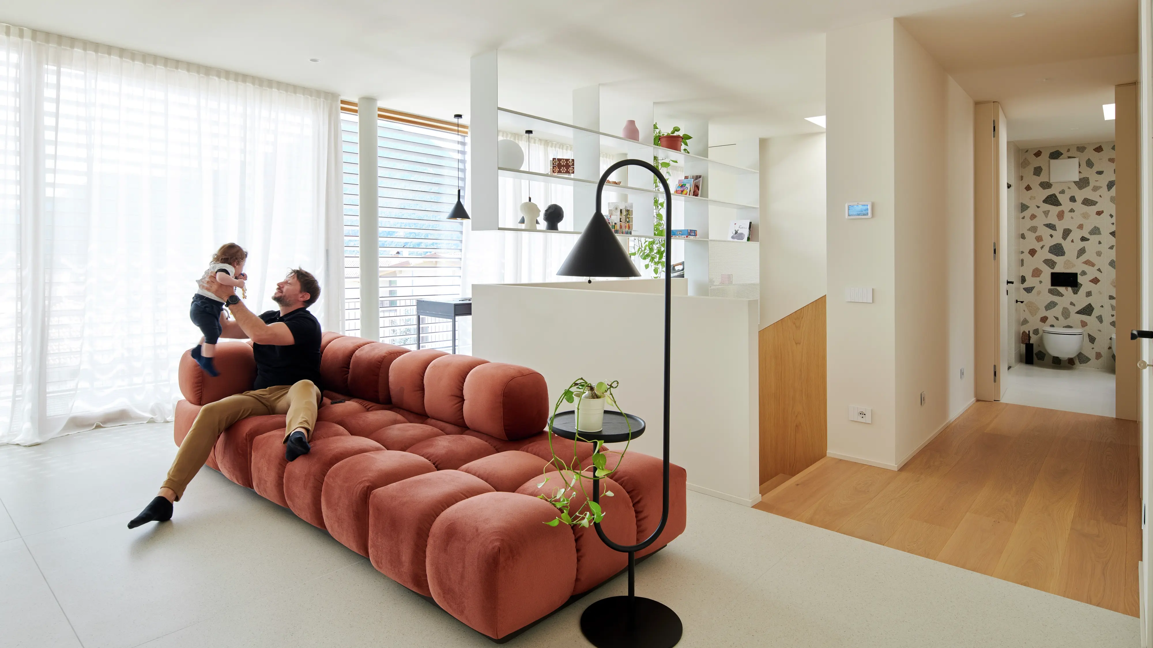 Salon moderne avec canapé en terre cuite, lampe noire, étagères ouvertes et salle de bain en terrazzo.