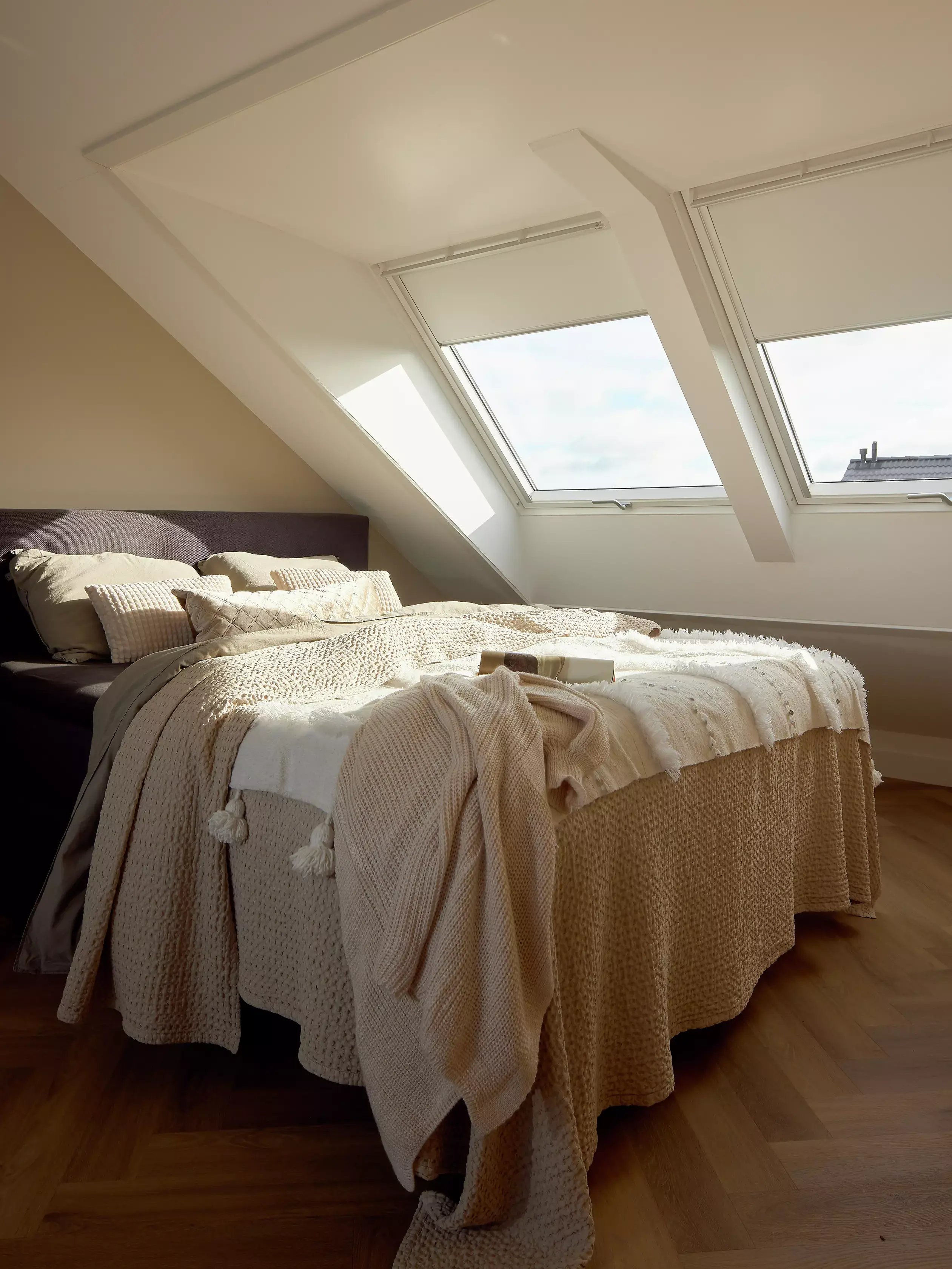Chambre sous combles éclairée par des lucarnes avec un lit douillet et des couvertures couleur crème.