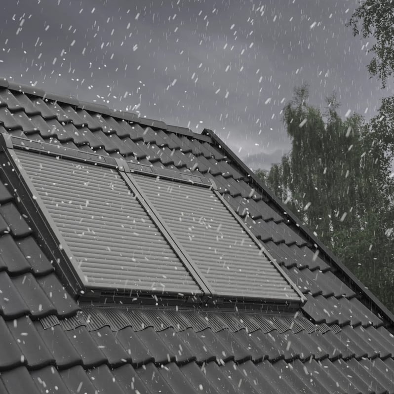 Tiled roof with closed VELUX roof window and solar roller shutter in weather with hail.