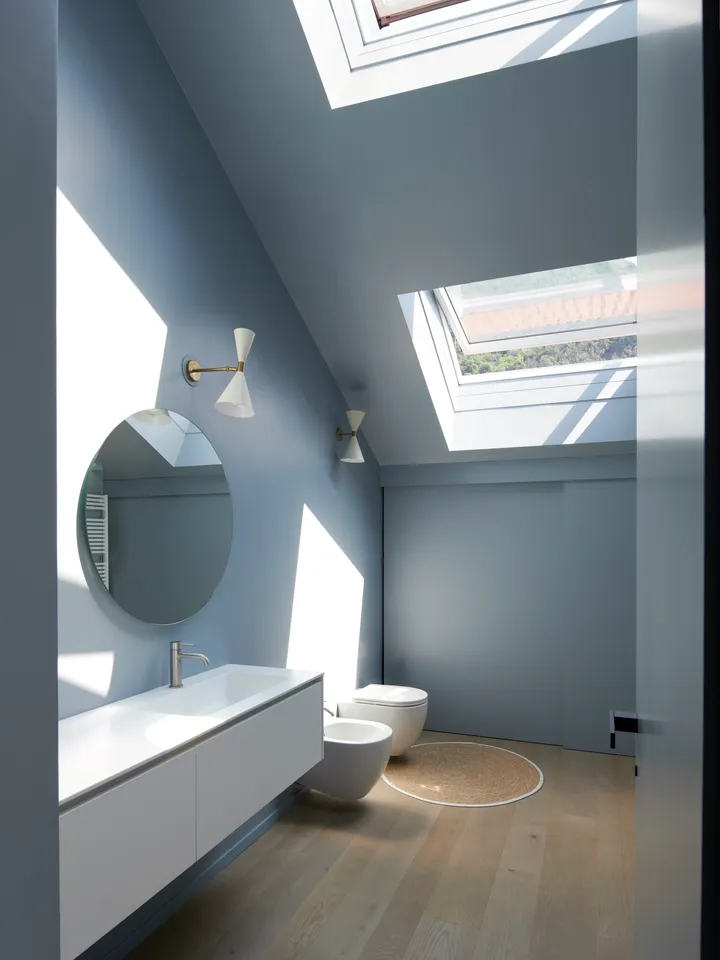  Gri-mavi duvarlar, açık ahşap zemin ve beyaz sıhhi tesisat malzemeleriyle minimalist bir banyo. Bir çatı penceresi, mekana doğal ışık girmesini sağlıyor.