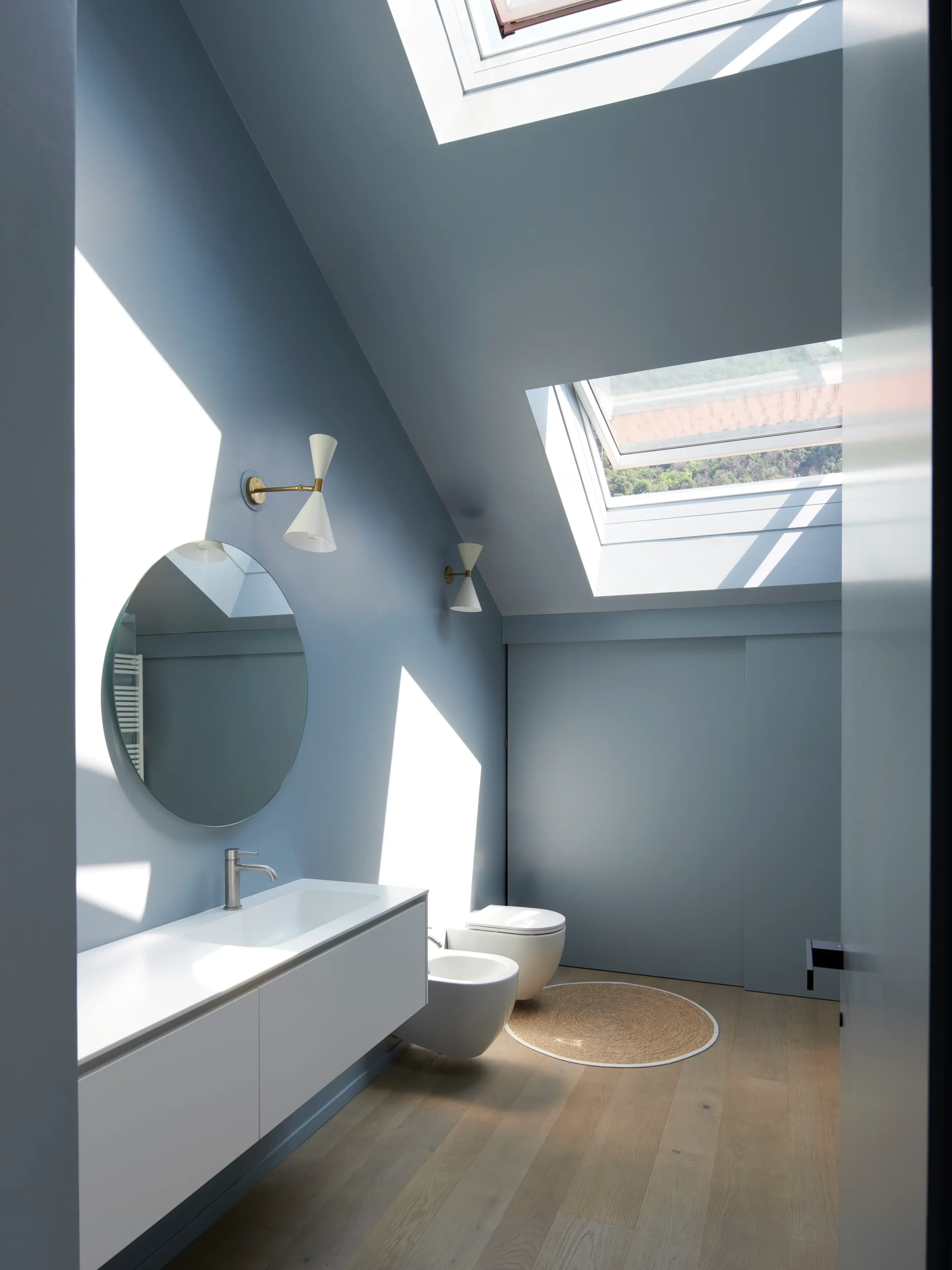 Une salle de bain minimaliste avec des murs gris-bleu, un plancher en bois clair, et des sanitaires blancs. Une fenêtre de toit projette la lumière naturelle sur l'espace.