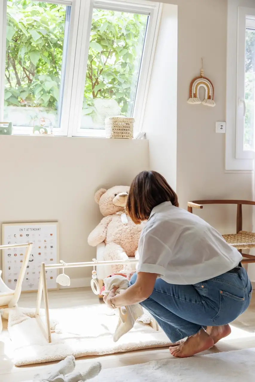 Une femme rangeant des jouets dans une chambre d'enfant lumineuse avec un grand ours en peluche, un portique de jeu et une lumière naturelle provenant des fenêtres de toit VELUX. 