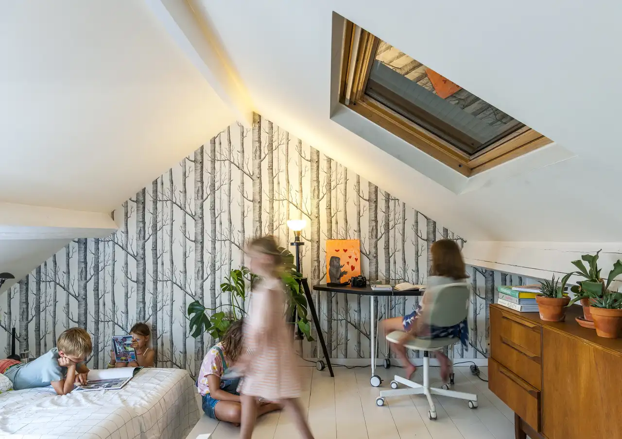 Des enfants jouent dans une chambre mansardée équipée d’une fenêtre de toit avec volet fermé.