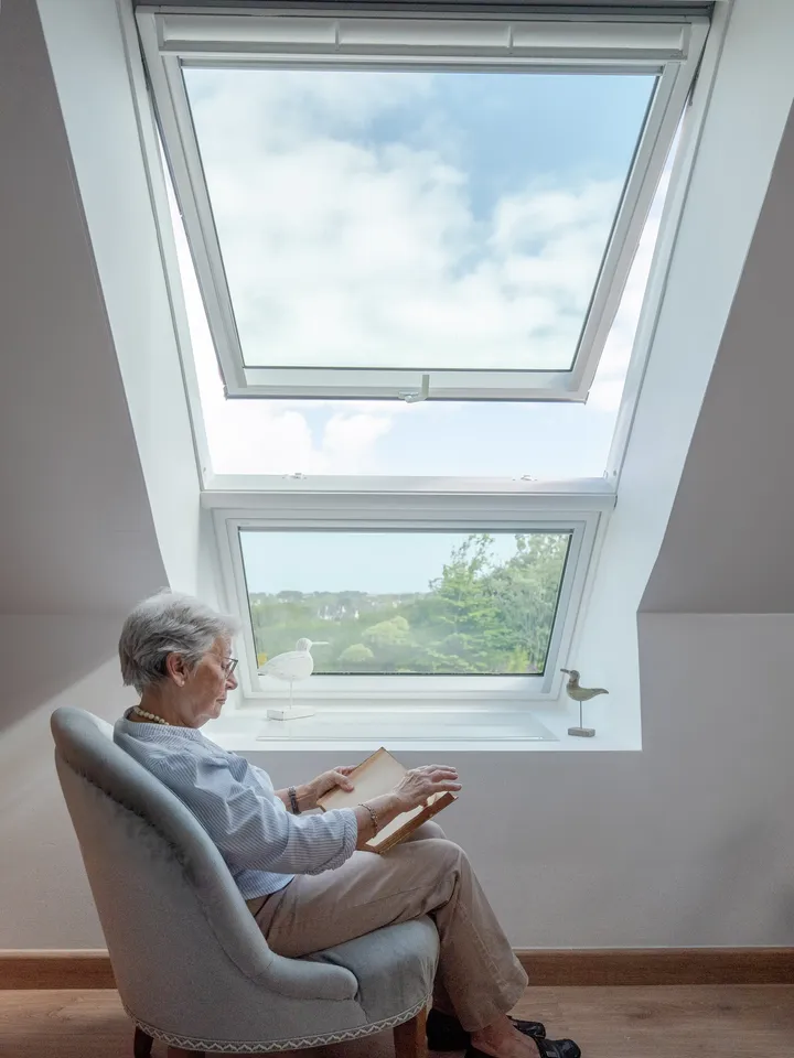 Gemütliche Leseecke auf dem Dachboden mit Sessel und geöffnetem VELUX Dachflächenfenster, das den blauen Himmel zeigt.