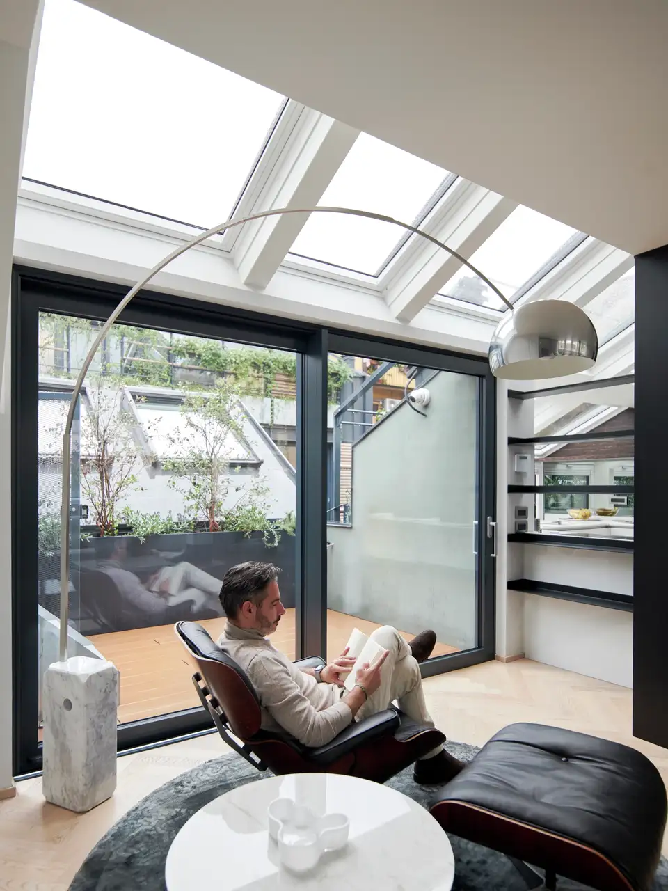 homme assis sur la chaise Eames Lounge lit un livre près des fenêtres de toit VELUX et lampe Arco