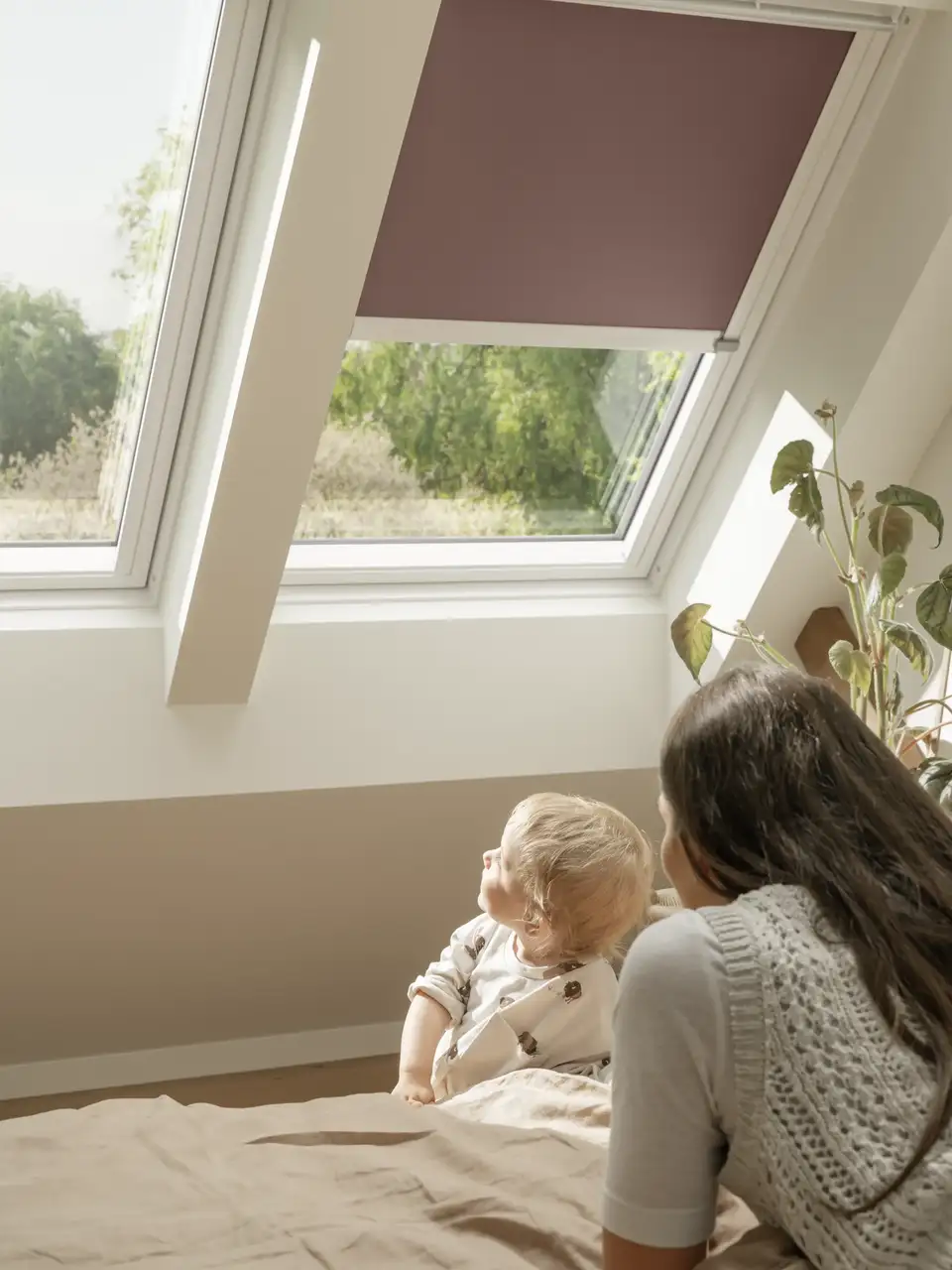 "Moeder met haar jonge kind op een tweepersoonsbed, kijkend door een VELUX dakvenster en natuurlijk zonlicht dat de kamer binnenvalt." 