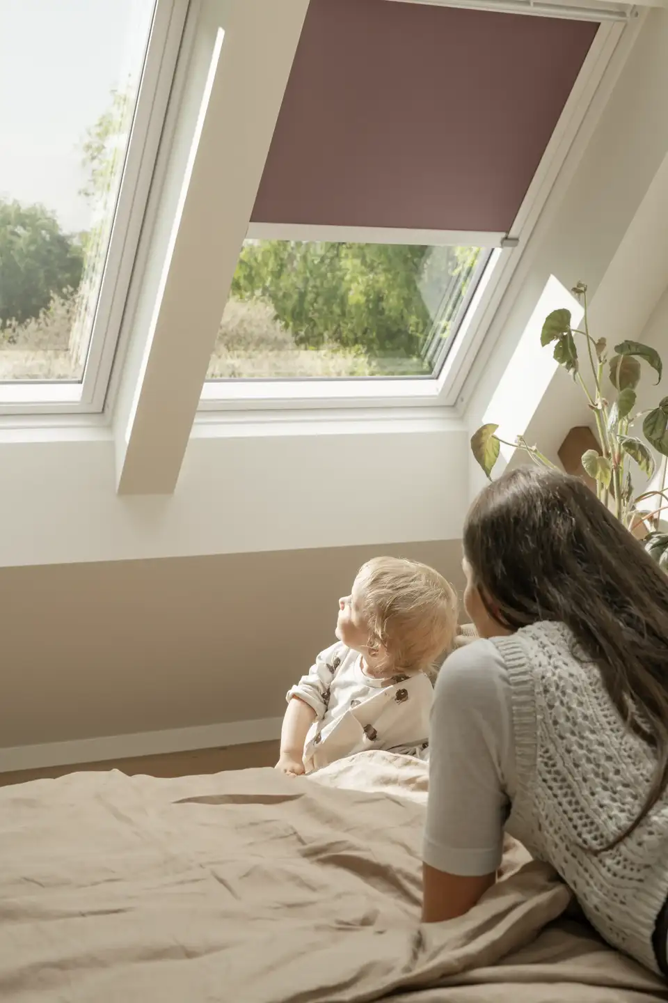"Mãe com o seu filho pequeno numa cama de casal a olhar por uma janela de telhado VELUX, com luz do sol natural a entrar pelo quarto." 