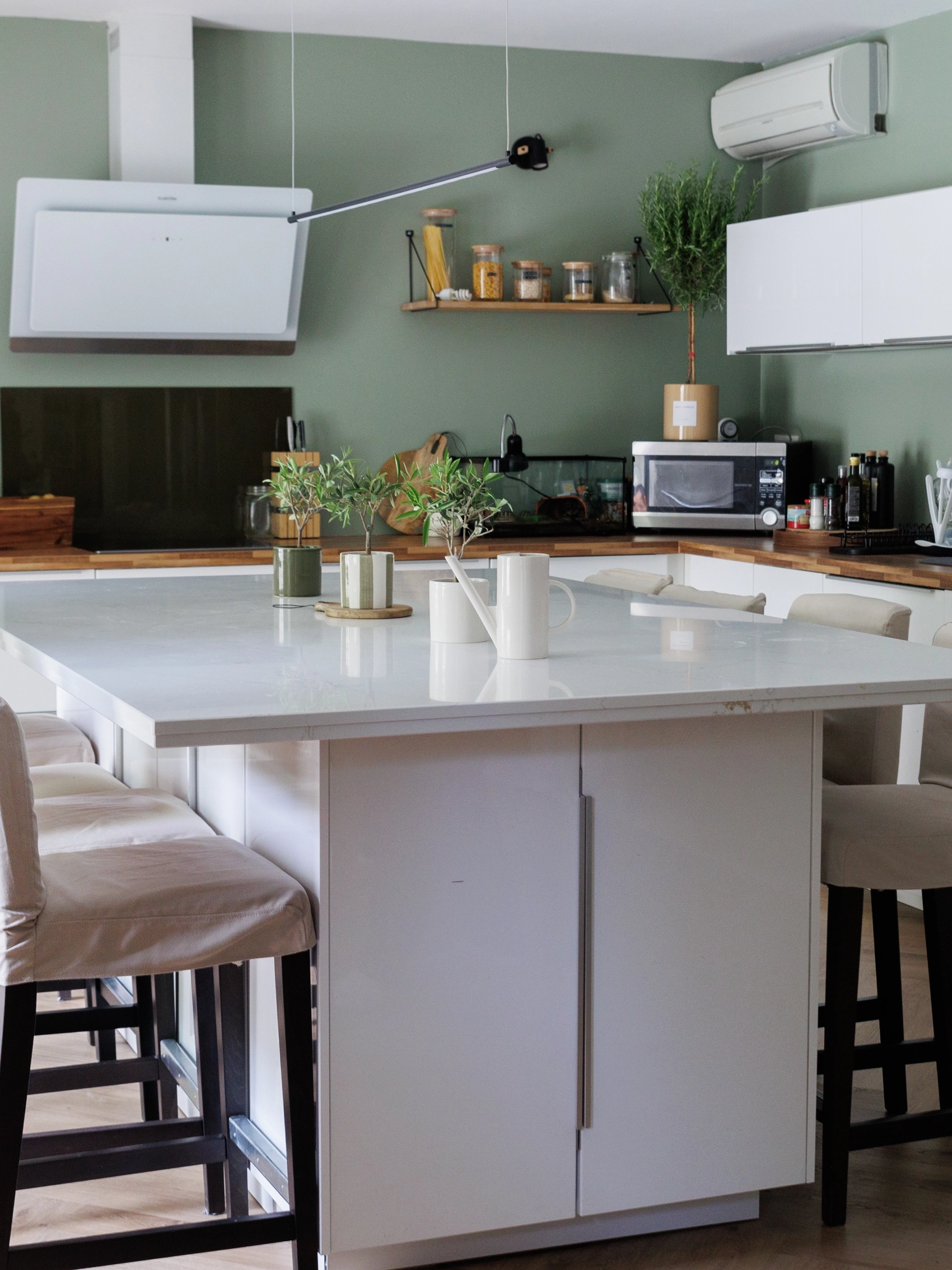 Cuisine moderne et fonctionnelle avec des armoires blanches, un îlot central avec des tabourets hauts, et des touches de décoration verte qui complètent le mur de couleur sage.