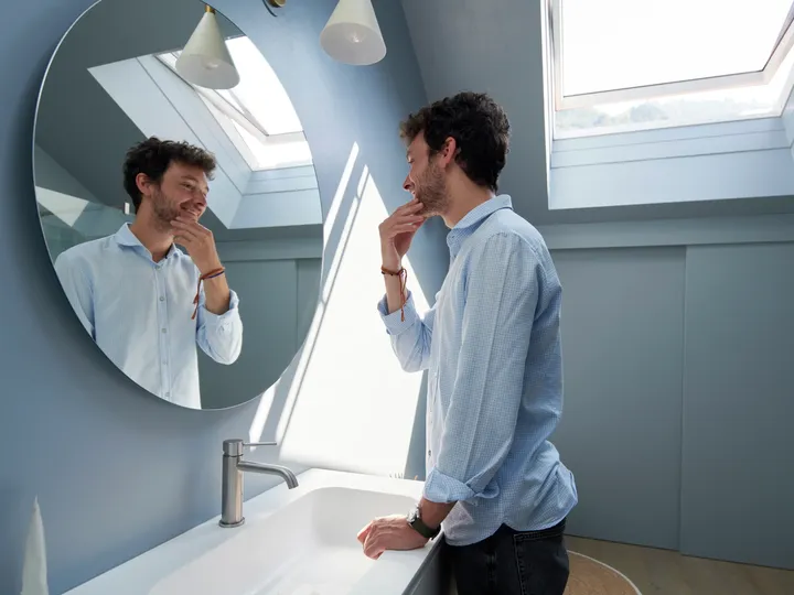 Modernes Badezimmer mit natürlichem Licht von VELUX Dachflächenfenster, rundem Spiegel und blauen Wänden.