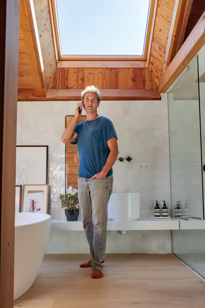 Une salle de bain lumineuse avec une baignoire blanche autoportante, de grandes fenêtres encadrées de bois avec vue sur la verdure, une fenêtre de toit et un comptoir minimaliste avec des décorations et des œuvres encadrées. 