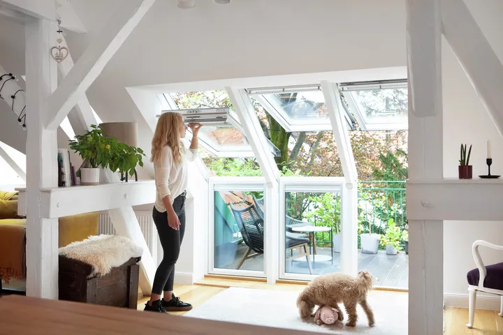 Frau öffnet Fenster ihres Dachbalkons im Wohnzimmer während ihr Hund mit Spielzeug spielt | VELUX Magazin