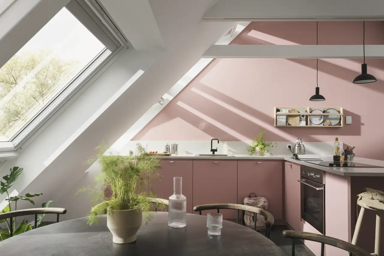 Cuisine mansardée éclairée par de grandes fenêtres de toit ; le mur vertical et les meubles de cuisine sont dans un rose doux. 