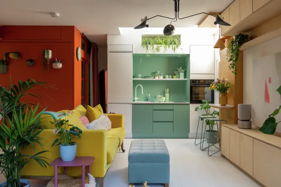 Salon moderne avec canapé jaune, cuisine vert d’eau et mur rouge brique, décoré de plantes et de meubles colorés dans un style années 80 revisité 