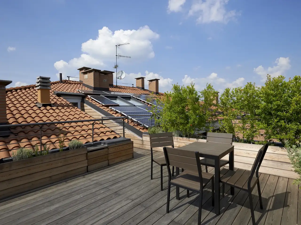  terrasse sur toit plat avec sol en lames de bois, table et chaises, jardinières et vue sur des toits en tuiles équipés de panneaux solaires 