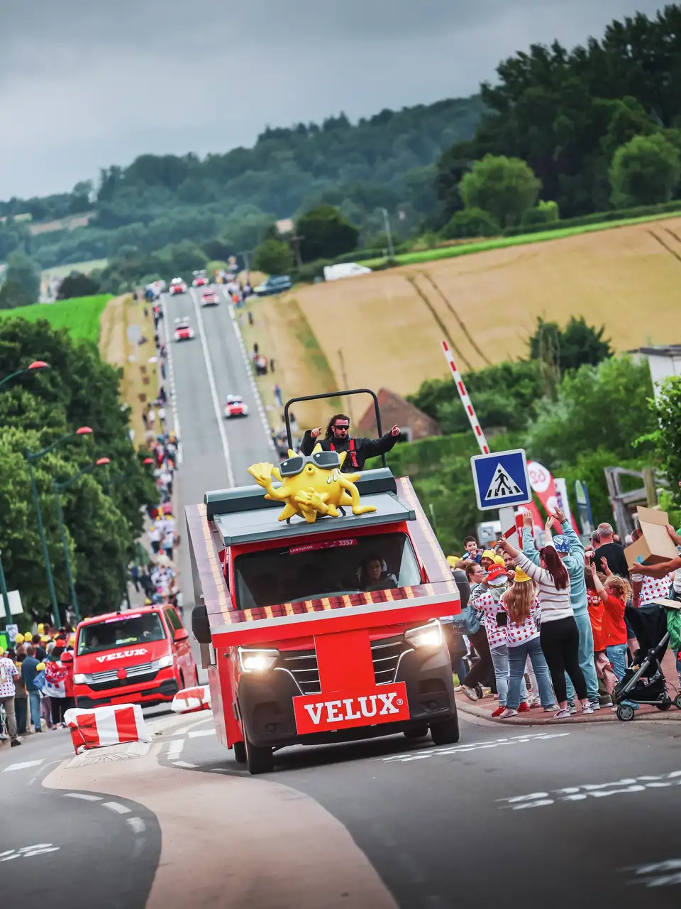 Vozilo VELUX v paradi Tour de France s kričečo množico in podeželskim ozadjem.