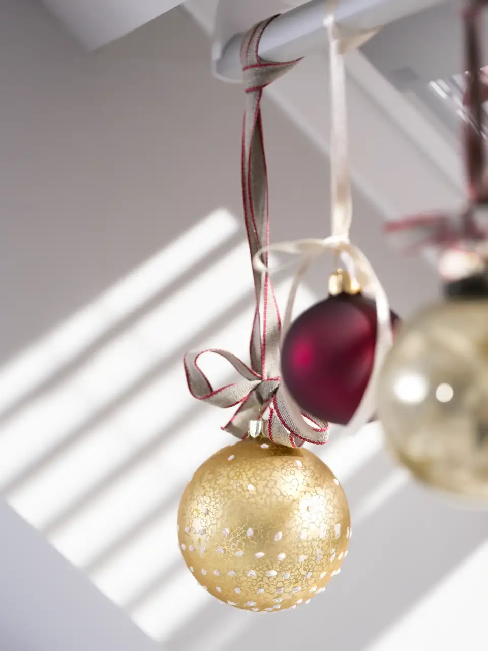 Palline natalizie rosse e dorate appese con nastri a una finestra per tetti VELUX, illuminate dalla luce naturale.