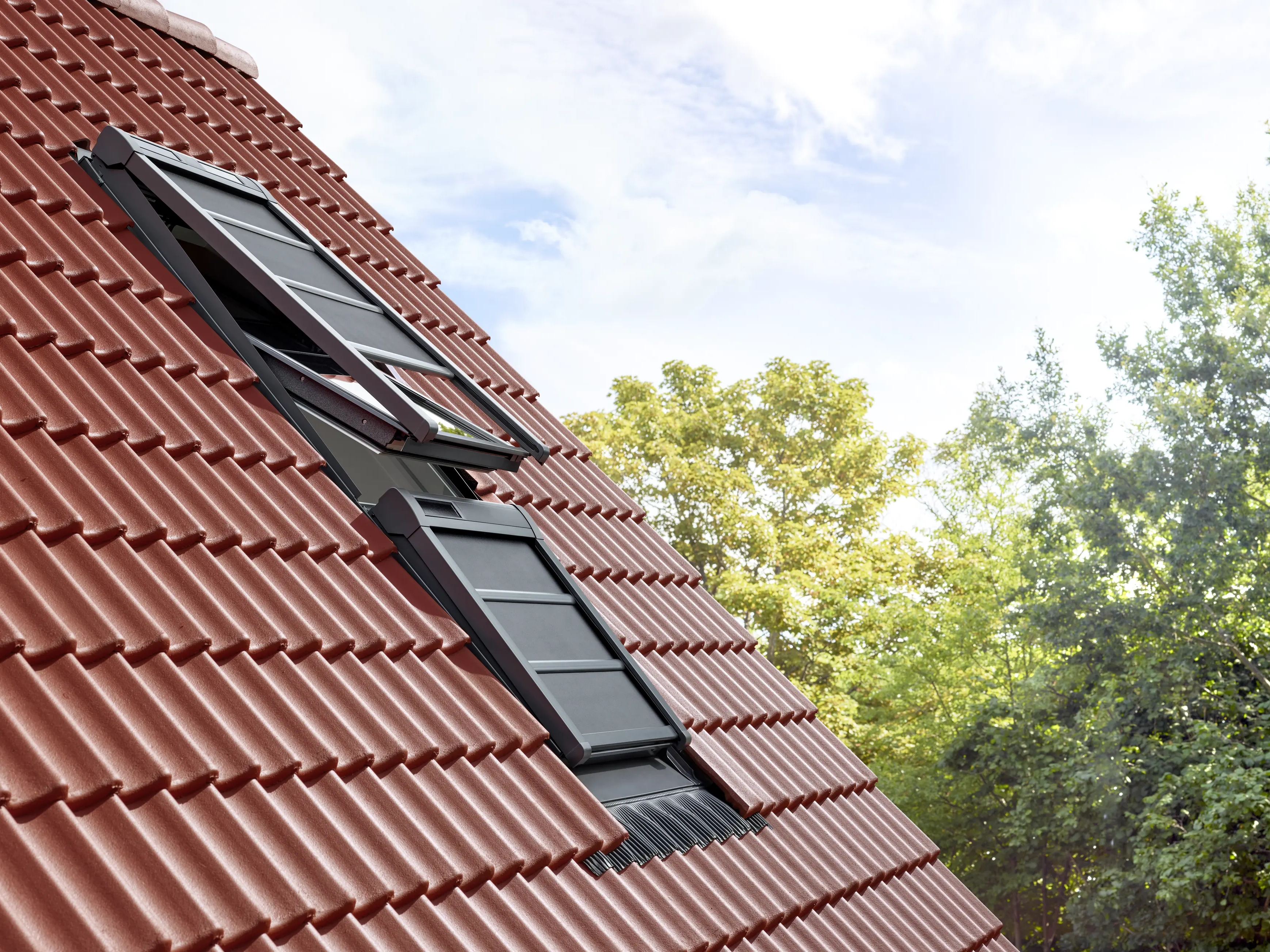 Toit en tuiles rouges à forte pente, avec une fenêtre de toit VELUX ouverte, entouré d’arbres.