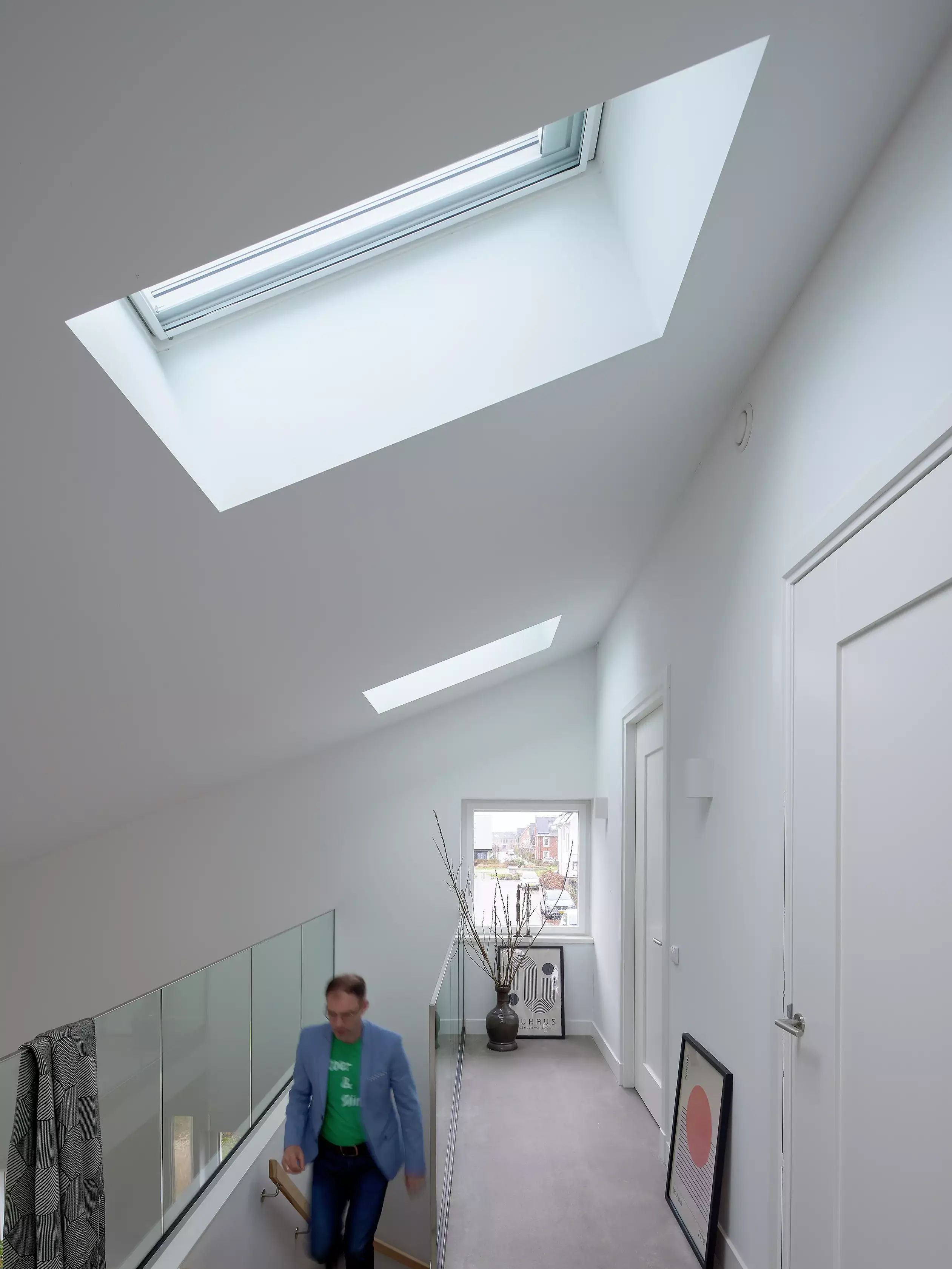 Couloir moderne avec lumière naturelle provenant d'une fenêtre de toit VELUX, balustrade en verre et décoration minimaliste.
