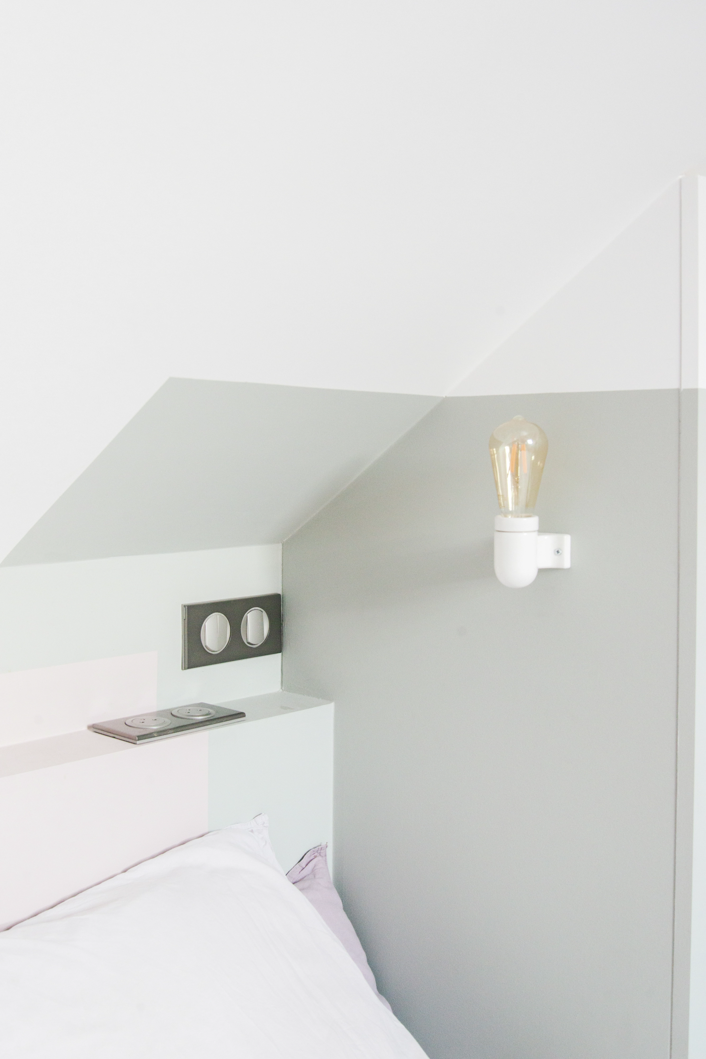 Minimālistisks bēniņu guļamistabas stūris ar slīpiem griestiem un sienas lampu.