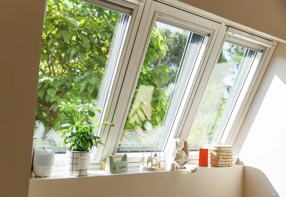 Trois fenêtres de toit VELUX au-dessus d'un rebord de fenêtre lumineux décoré avec des plantes, des jouets et des petits paniers, donnant sur une verdure luxuriante. 