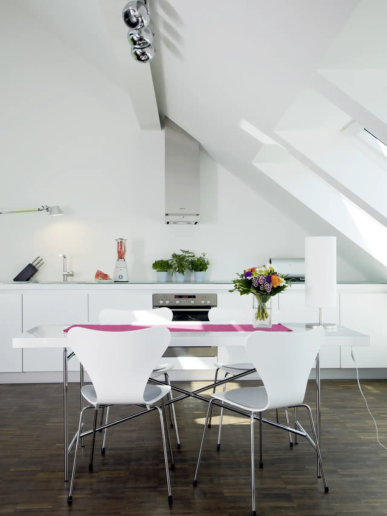 Cuisine blanche avec table et chaises blanches aux pieds métalliques, fenêtres de toit et spots en métal.