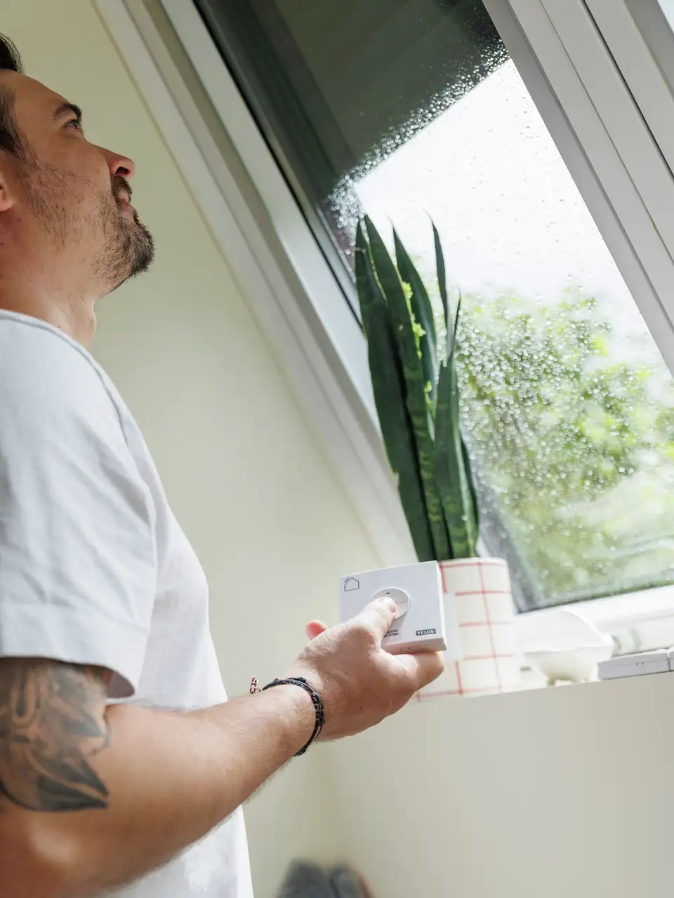 Persoon gebruikt afstandsbediening voor VELUX dakraam, regenachtig uitzicht buiten.