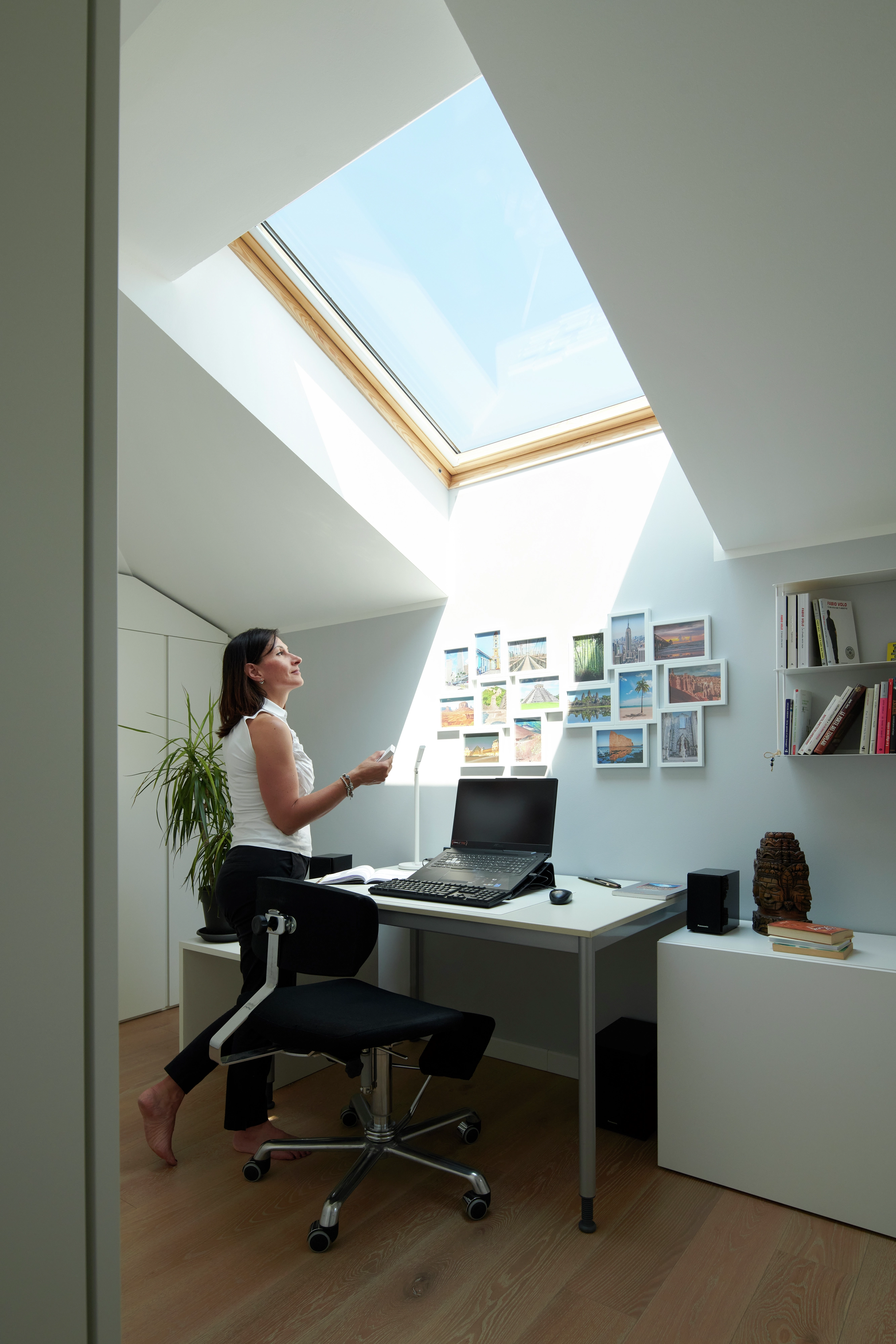 Bureau à domicile dans le comble avec fenêtre de toit VELUX, bureau blanc, ordinateur portable et photos murales.