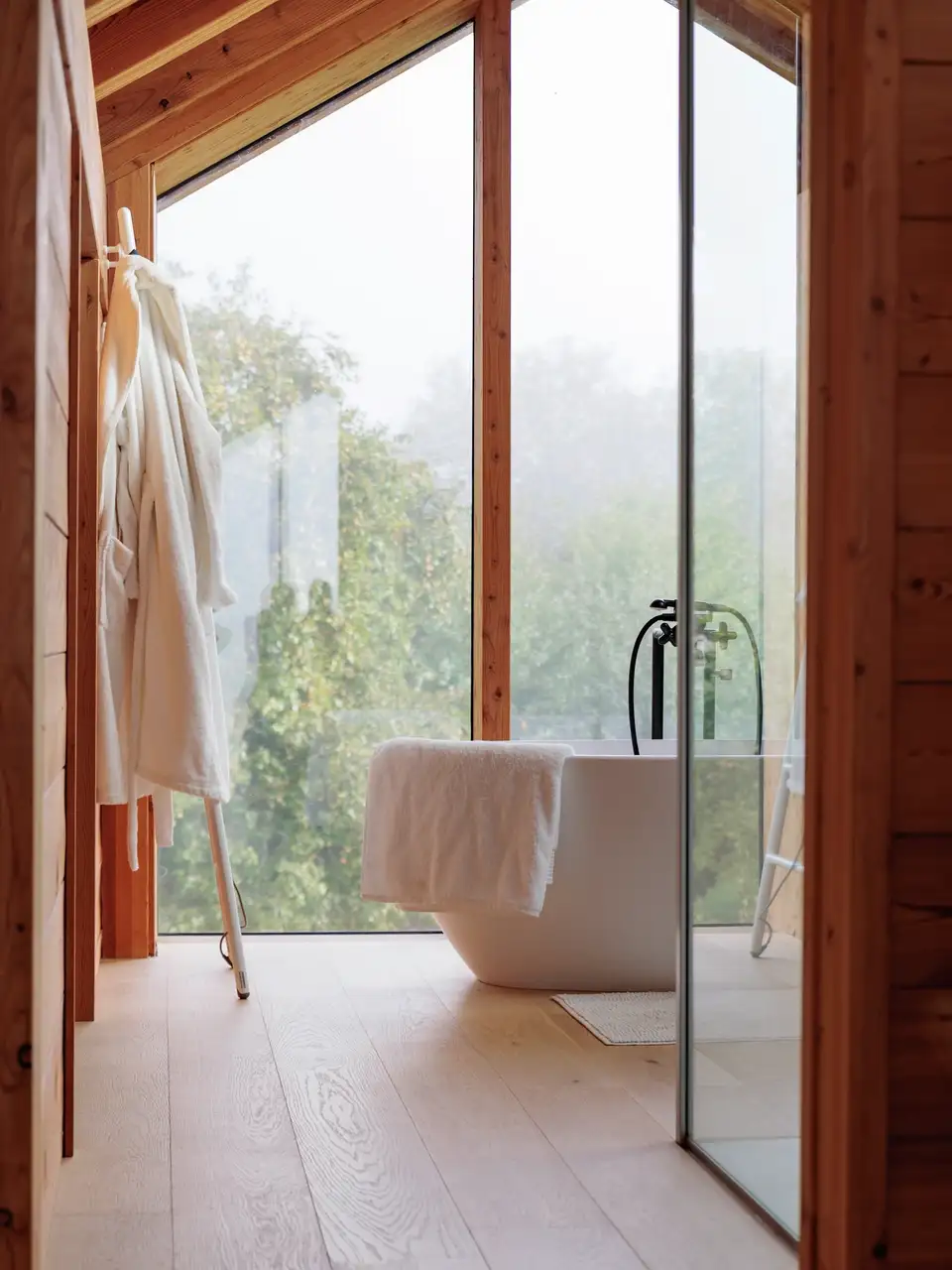 Een badkamer met veel natuurlijk licht, houten wandbekleding, een lichte vloer, een ruim vrijstaand bad en een glazen deur die uitzicht biedt op een groene omgeving.