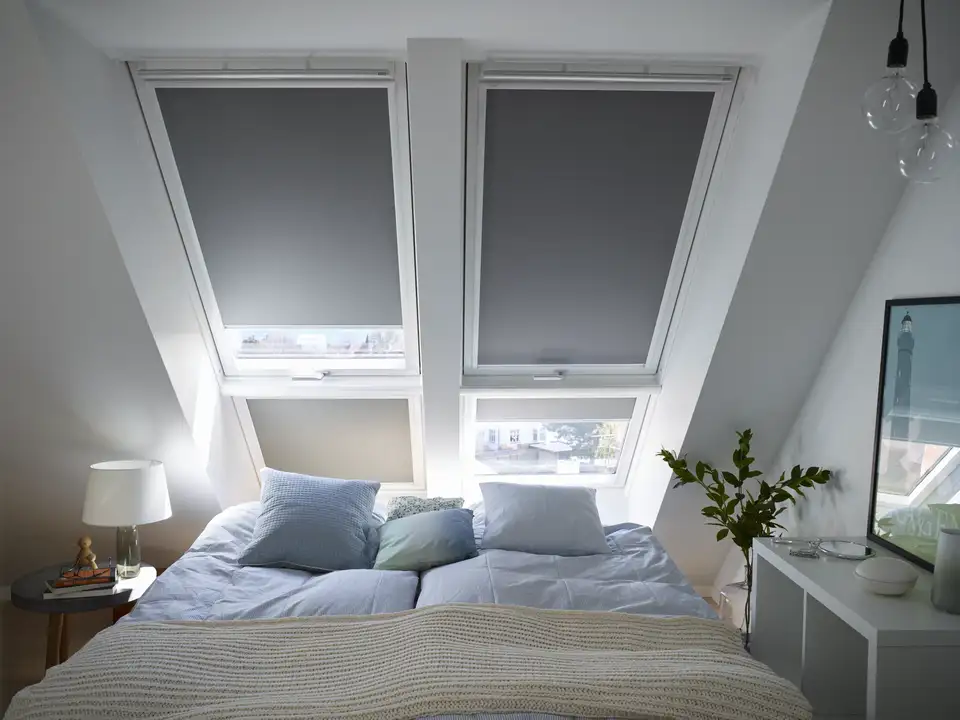 Een goed verlichte, moderne slaapkamer met een schuin plafond, met twee dakramen die natuurlijk licht door de ruimte laten stromen. Het bed is opgemaakt met wit en lichtblauw beddengoed en er staan nachtkastjes met lampen aan weerszijden. Een plant voegt een vleugje groen toe aan de kamer.