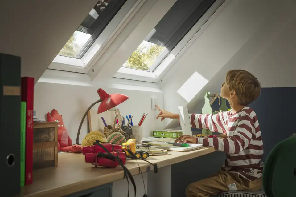 Een kind dat aan zijn bureau zit, opent het VELUX rolluik met behulp van een afstandsbediening. 