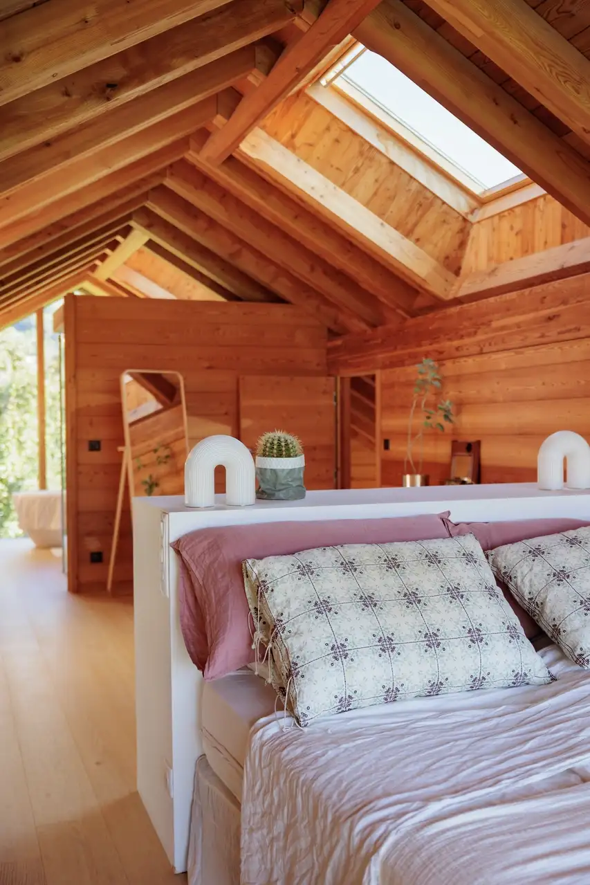 Une chambre loft chaleureuse en bois avec des poutres apparentes, des textiles doux et une fenêtre de toit VELUX laissant entrer la lumière naturelle.