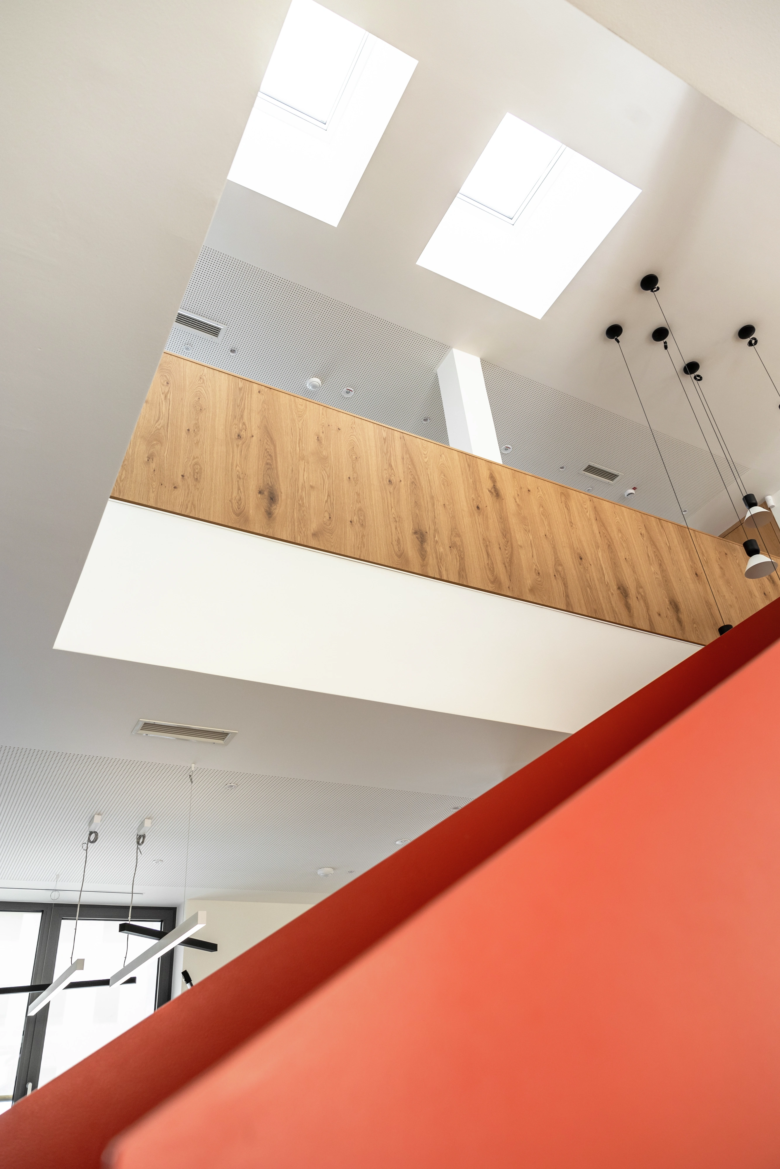 Moderní interiér s vysokým stropem, střešními okny VELUX, dřevěným balkonem a červeným schodištěm