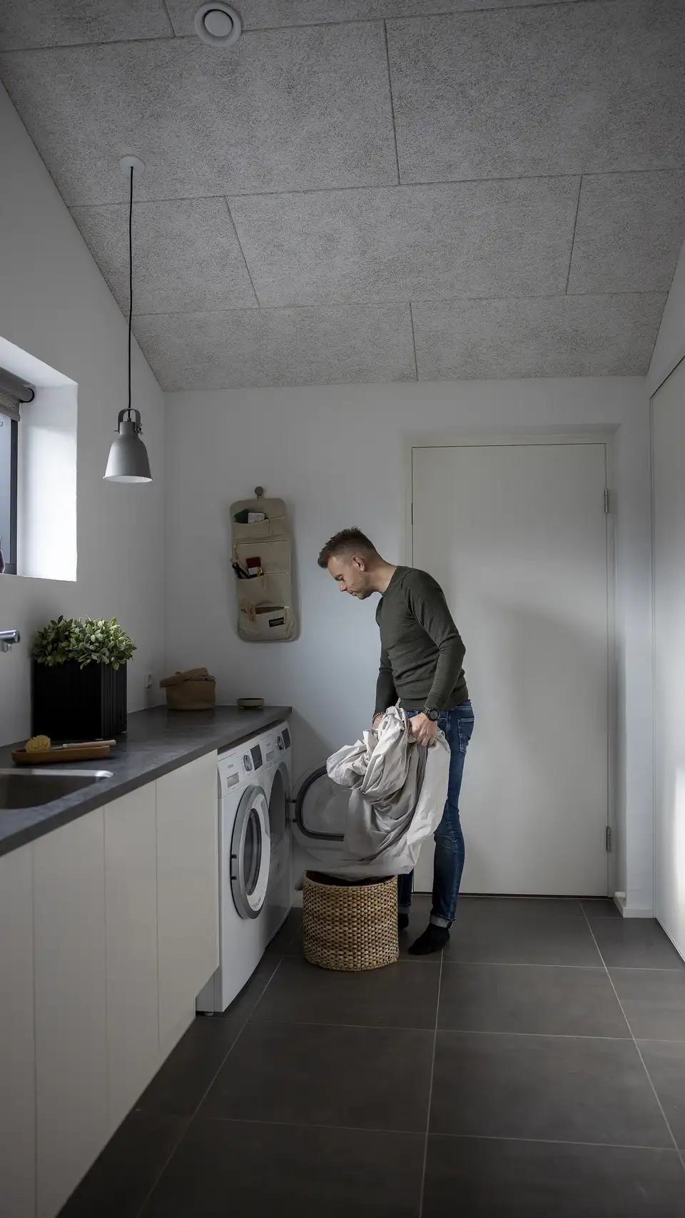 homme déposant du linge dans une panière, dans une petite buanderie avec lave-linge sous plan de travail et fenêtre latérale