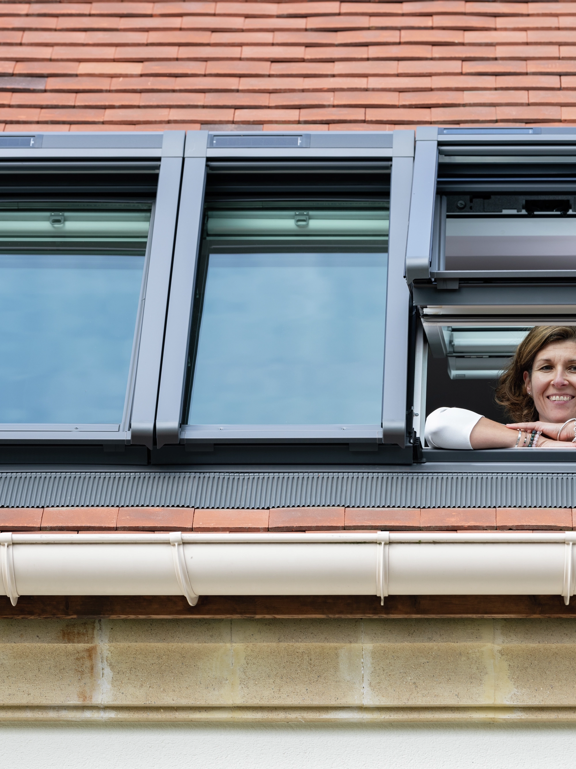 Une femme devant une fenêtre de toit VELUX ouverte sur un toit en tuiles, offrant une vue sur le ciel.