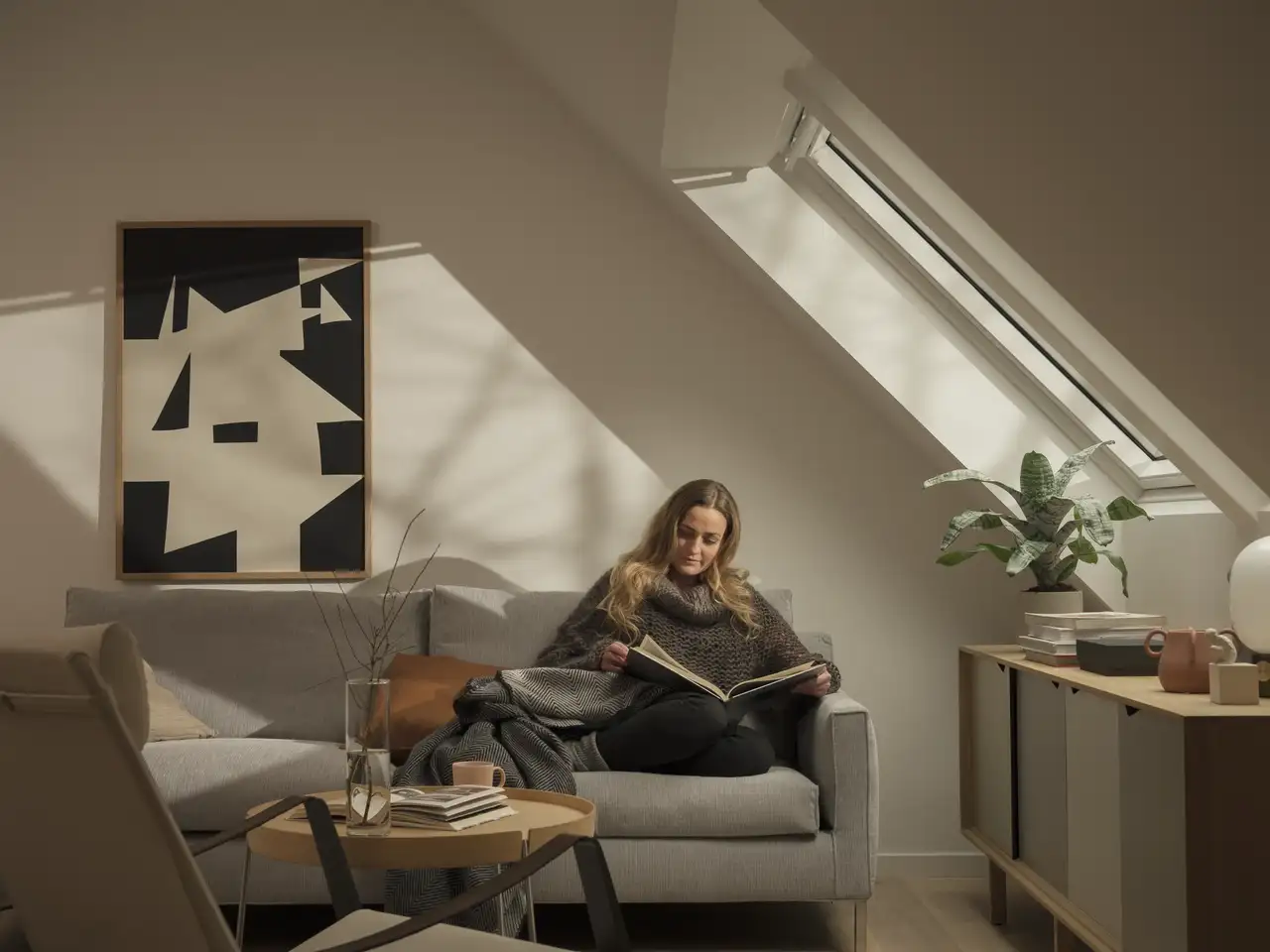 Frau sitzt eingekuschelt auf Couch unter Dachfenster und liest ein Buch | VELUX Magazin