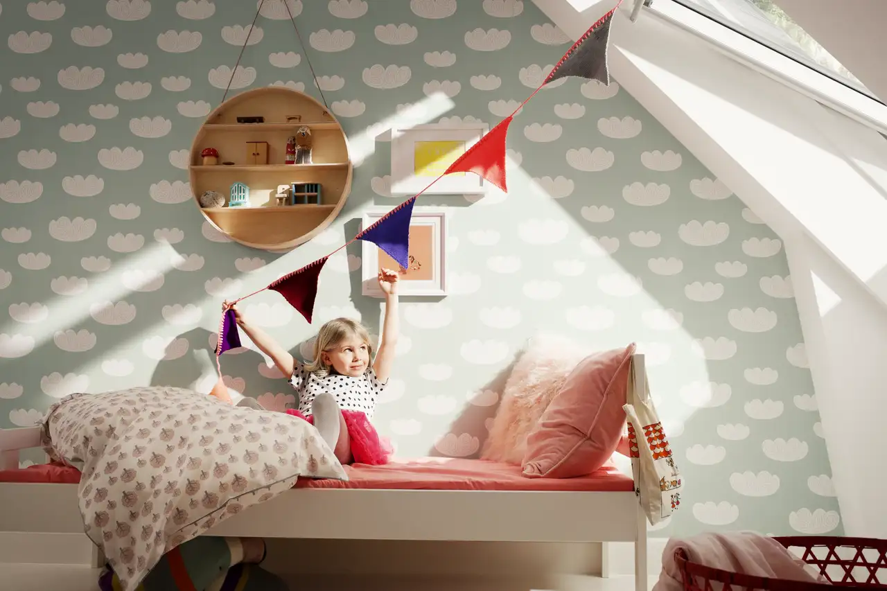 Mädchen sitzt auf Bett im Sonnenlicht in einem Kinderzimmer im Danish-Pastel-Stil mit pastelligen Farben, Dachfenster und Wimpelkette | VELUX Magazin