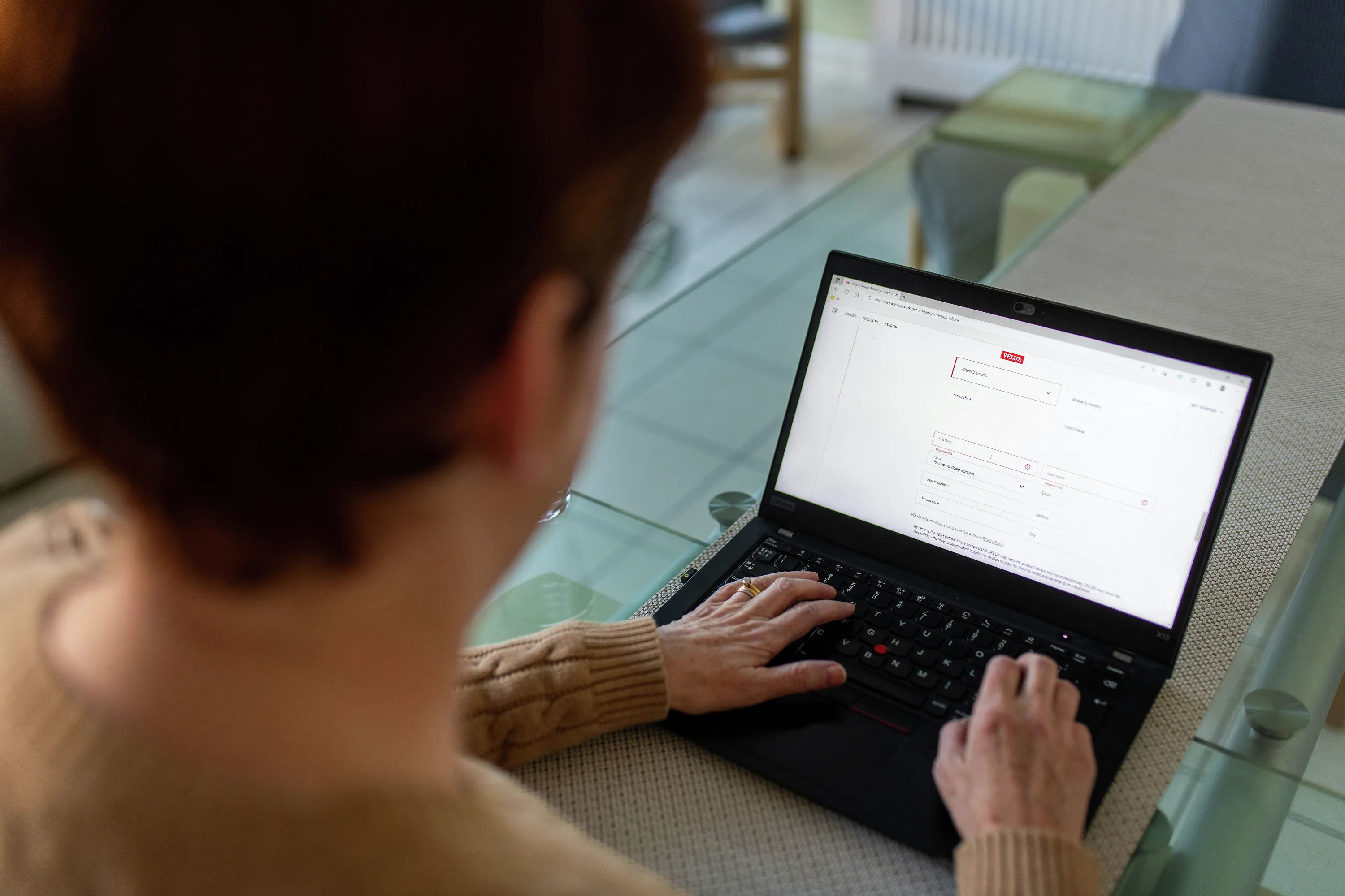 Woman typing on laptop at glass table, focused on screen.