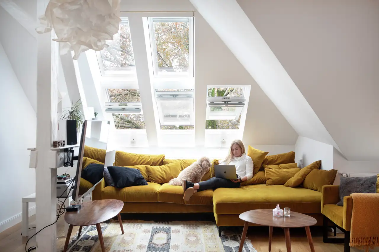 Woman sitting on a sofa with a laptop, accompanied by a dog, under roof windows letting in natural light. | VELUX