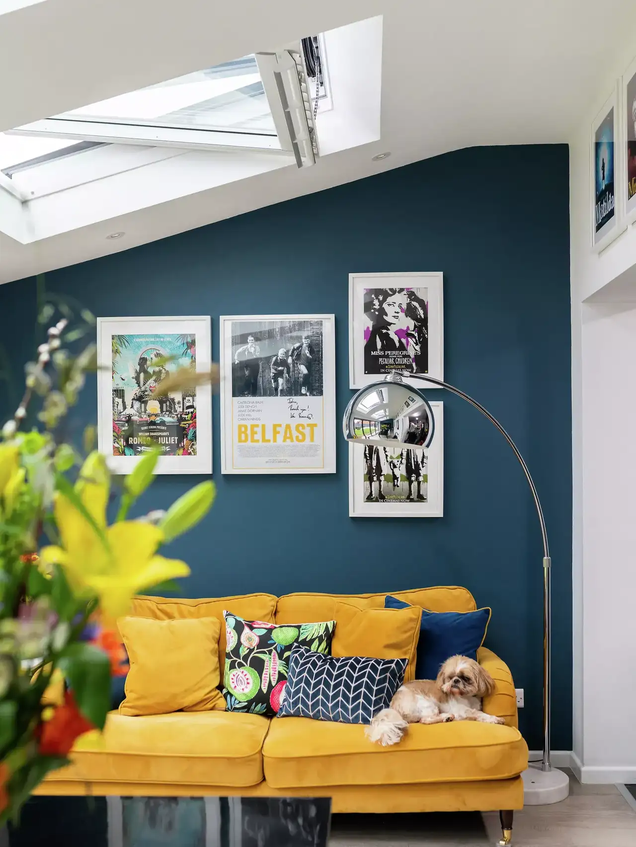 Gelbes Sofa mit Kissen, blaue Wand mit Kunst, VELUX Dachflächenfenster und Stehlampe im Wohnzimmer.