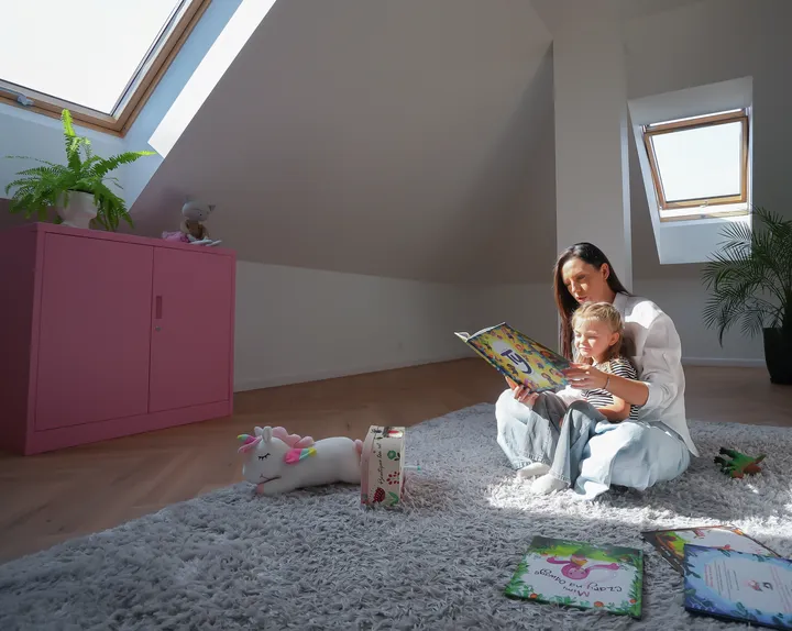 Mama czytająca dziecku w jasnym pokoju na poddaszu z oknami dachowymi | VELUX