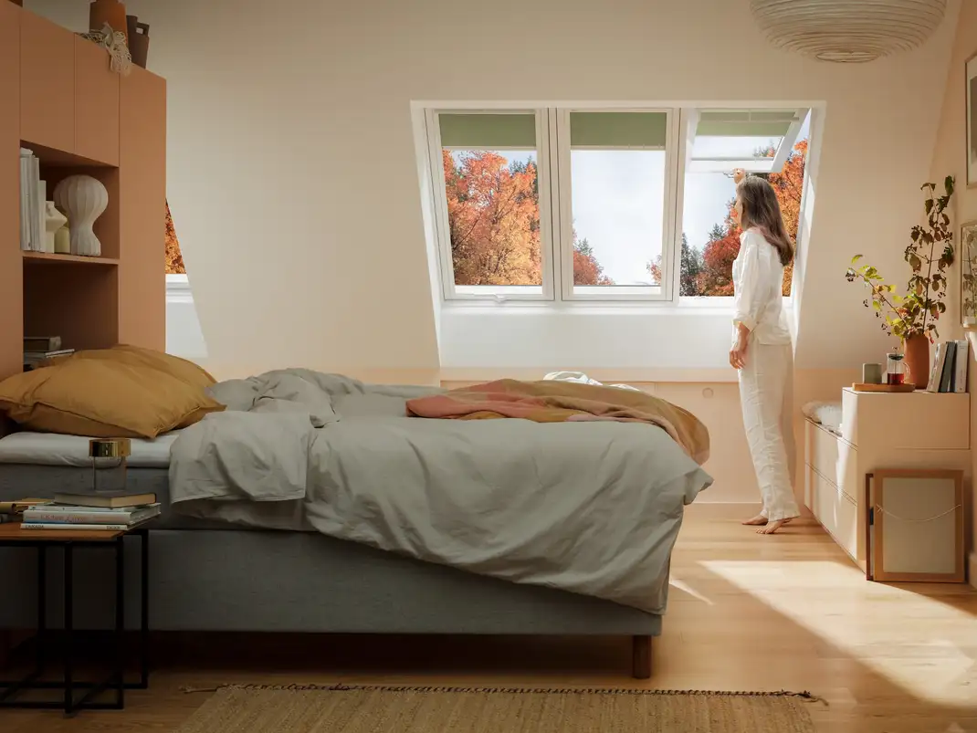 Attic bedroom with sloped ceiling and VELUX roof window, woman enjoying view.