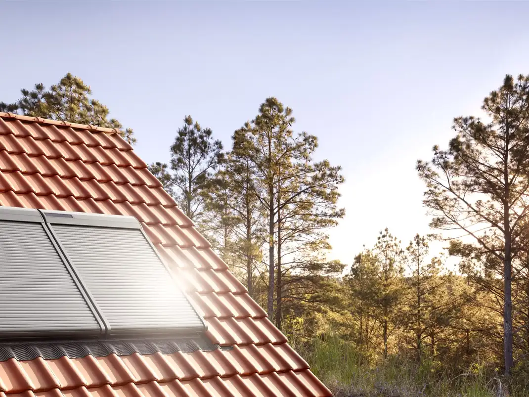 Red tiled roof with closed VELUX window and pine trees in background.
